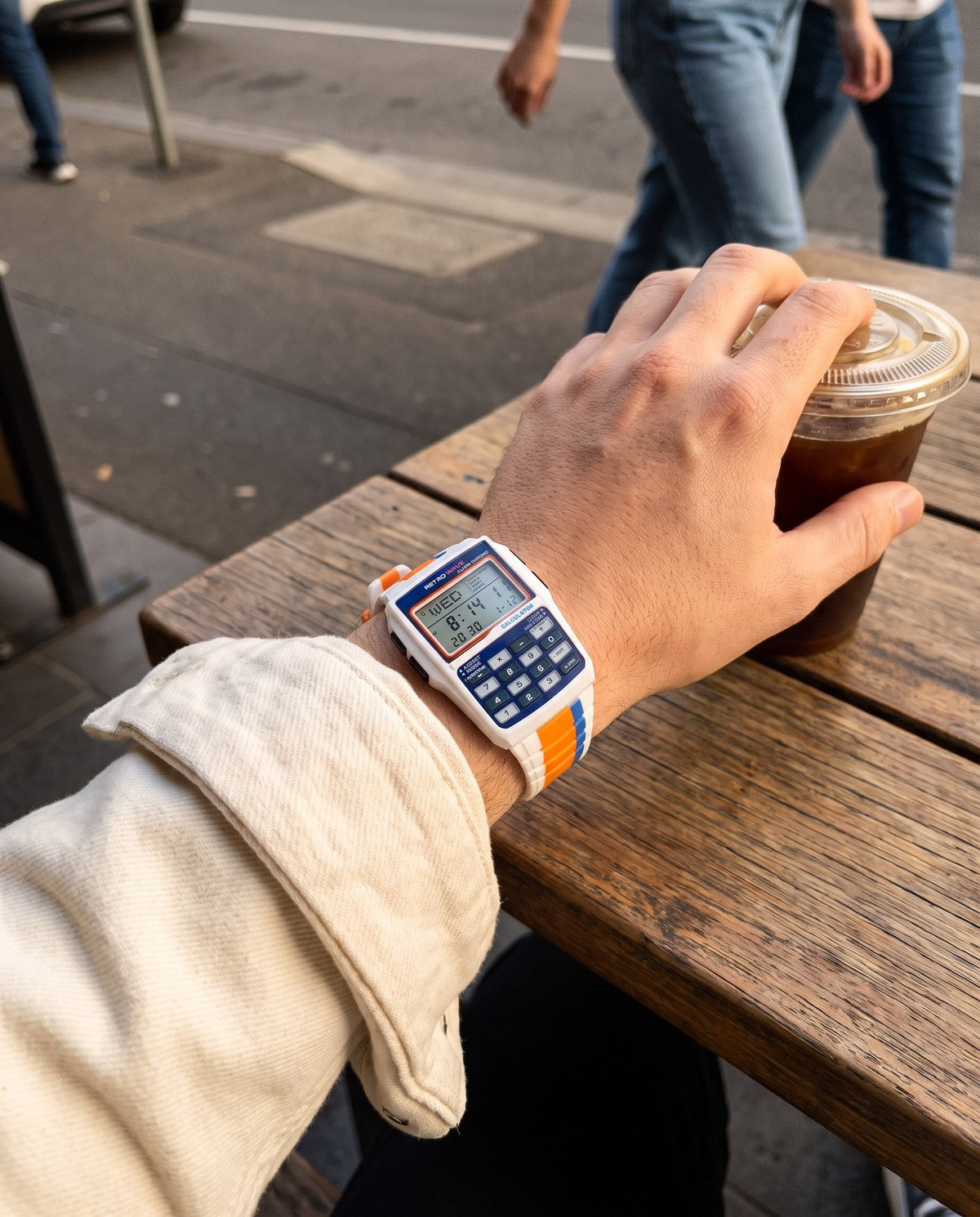 Left hand holding an iced coffee at a cafe table, wearing the Blue RetroWave calculator watch